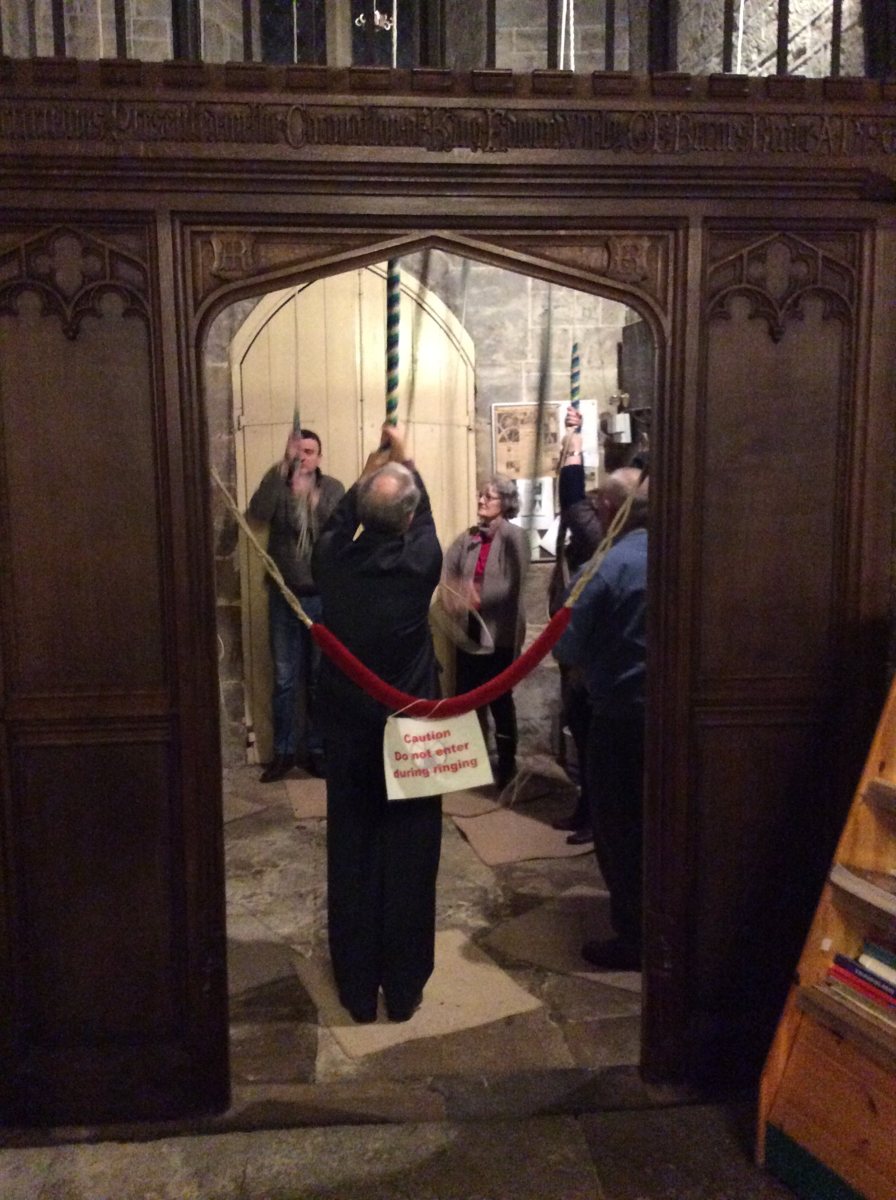 Bell ringers holding their ropes in the ringing chamber at the foot of the tower, seen through a carved wooden screen with a rope barrier and a sign saying Do not enter during ringing