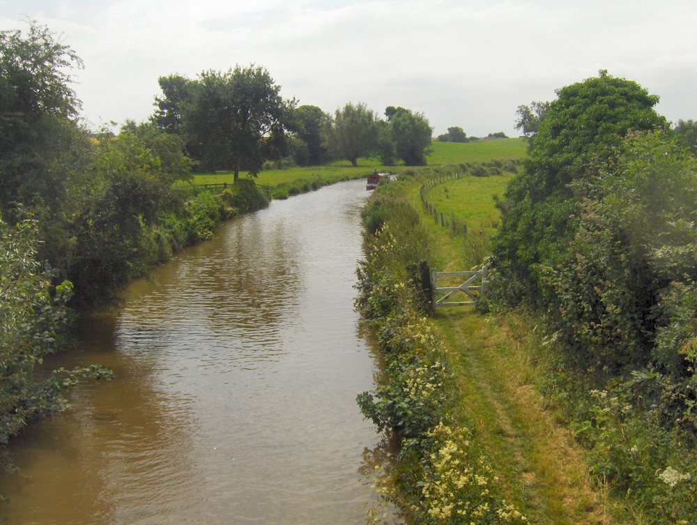 The Oxford Canal running between hedges and meadows, with the grassy towpath and a white gate on the right and a narrowboat in the distance
