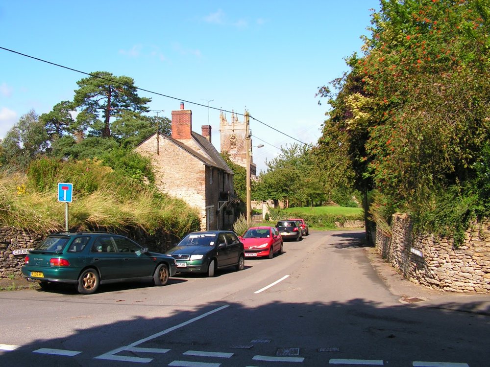 The corner of Church Street with a stone cottage and parked cars, and the church tower behind