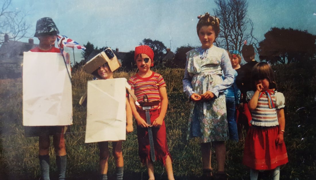 Children in fancy dress at the fete, including a pirate with a sword and eye patch, a girl in hair curlers and a housecoat, and two children in cardboard costumes
