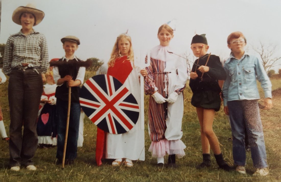 Six children in fancy dress standing in a field, including Britannia with a Union Jack shield and trident, a clown, a chimney sweep and Robin Hood