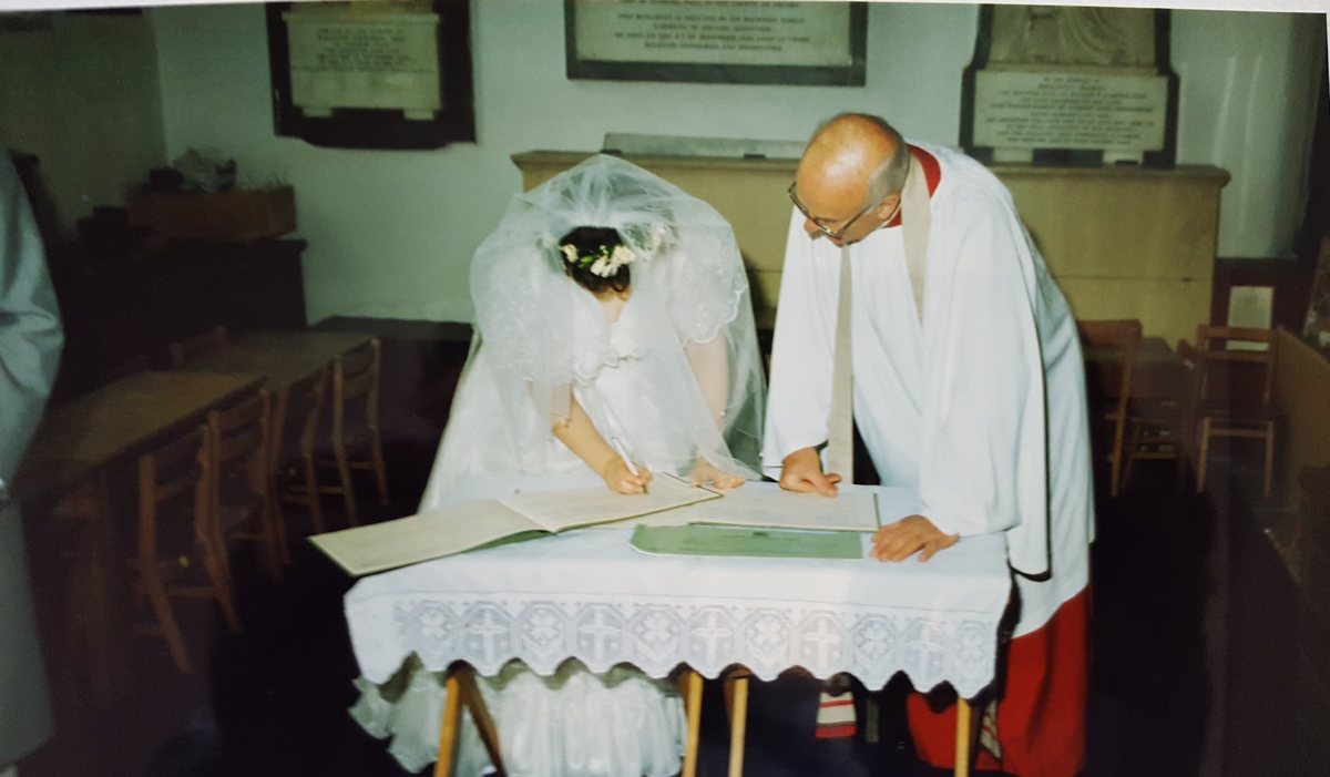 Canon Hewitt Wilson in surplice and red cassock beside a bride in a veil signing the marriage register at a table in St James's