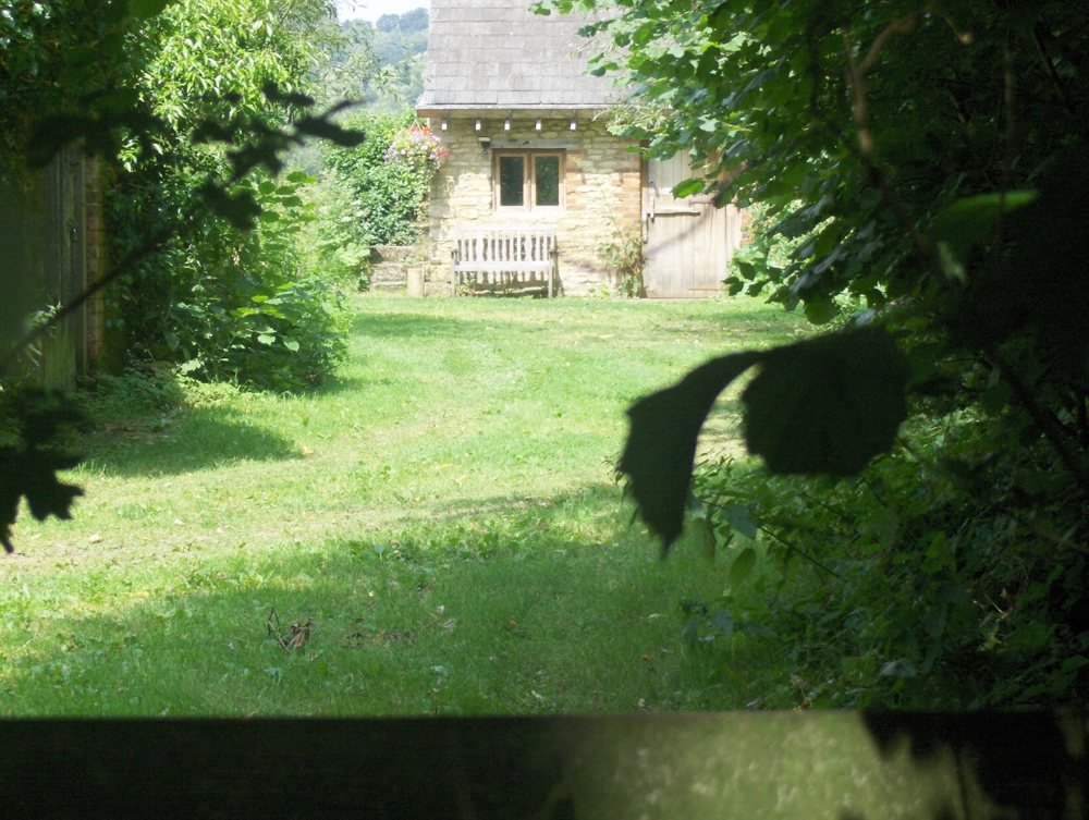 A small stone cottage with a bench and a wooden door, seen across a lawn through trees